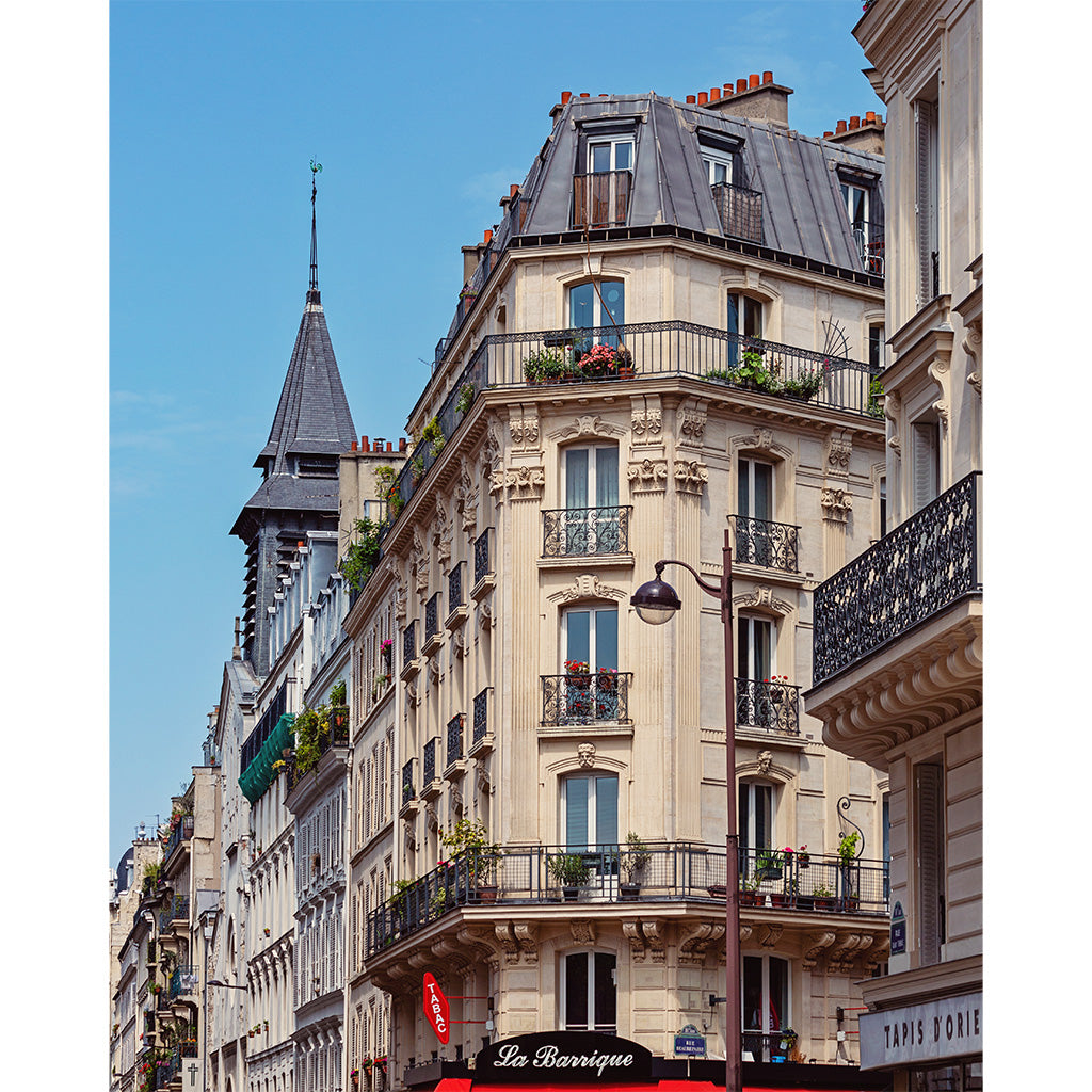 A classic Parisian-style apartment building facade with ornate cream stucco walls, black wrought-iron balconies, potted plants, and a dark gray slate roof under a clear blue sky.